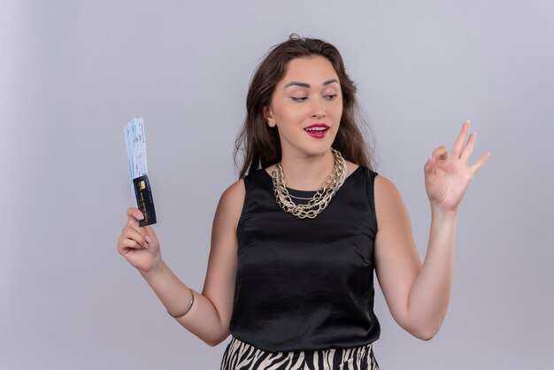 Souriante jeune femme voyageur vêtu d'un maillot noir tenant des billets et montre un geste okey sur un mur blanc