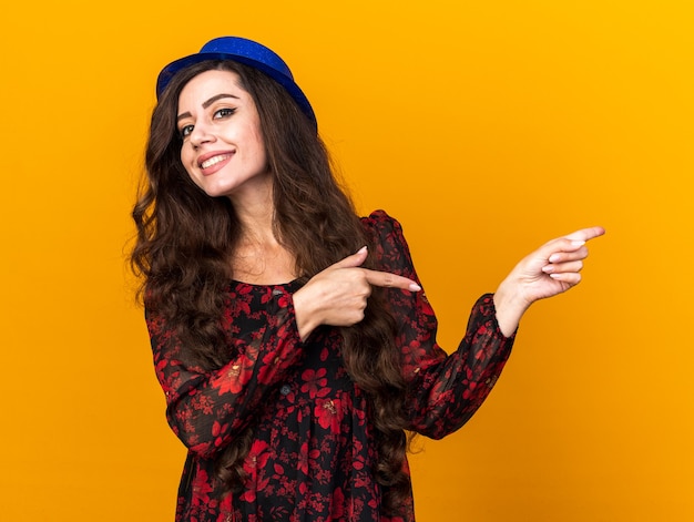 Souriante jeune fêtarde portant un chapeau de fête pointant sur le côté isolé sur un mur orange