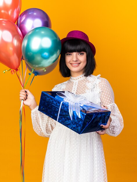 Souriante jeune fêtarde portant un chapeau de fête tenant des ballons et étirant un paquet cadeau isolé sur un mur orange