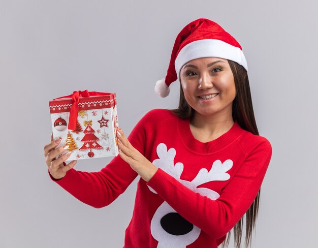 Souriante jeune fille asiatique portant un chapeau de Noël avec chandail tenant une boîte-cadeau isolé sur fond blanc