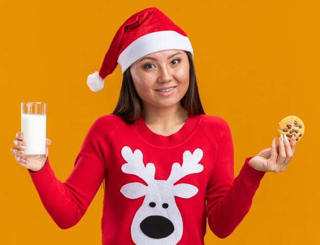 Souriante jeune fille asiatique portant chapeau de Noël avec chandail tenant un verre de lait avec des biscuits isolés sur fond orange