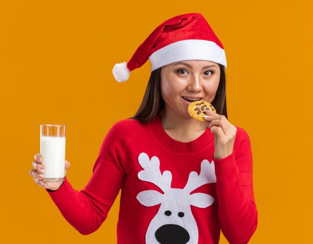 Souriante jeune fille asiatique portant un chapeau de Noël avec chandail tenant un verre de lait en essayant des cookies isolés sur un mur orange