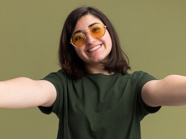 Souriante jeune fille assez caucasienne dans des lunettes de soleil fait semblant de tenir la caméra prenant selfie sur vert olive