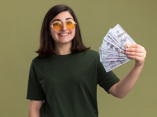 Souriante jeune fille assez caucasienne à lunettes de soleil détient de l'argent sur vert olive