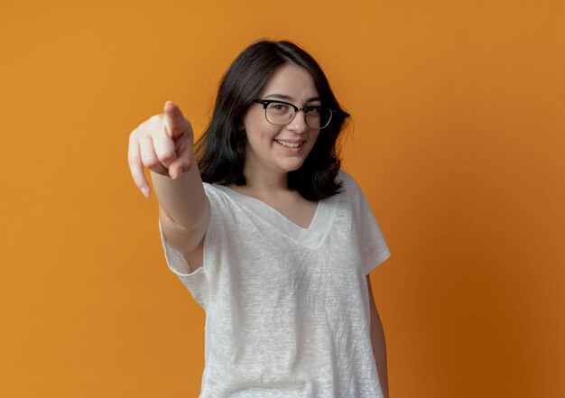 Souriante jeune fille assez caucasienne portant des lunettes pointant sur la caméra isolée sur fond orange avec espace copie