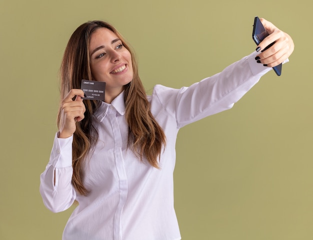 Souriante jeune fille assez caucasienne tient la carte de crédit et regarde le téléphone prenant selfie sur vert olive