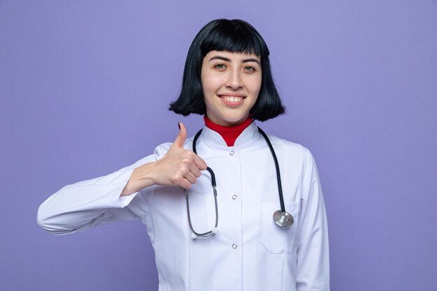 Souriante jeune fille assez caucasienne en uniforme de médecin avec stéthoscope pouce vers le haut