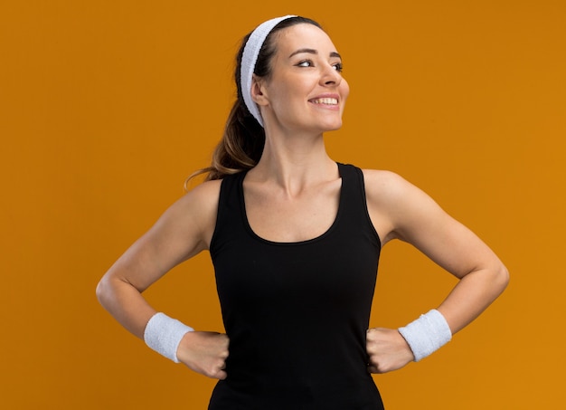 Souriante jeune fille assez sportive portant un bandeau et des bracelets gardant les mains sur la taille en regardant le côté isolé sur le mur orange