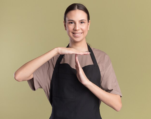 Souriante jeune fille de barbier brune en gestes uniformes signe de délai d'attente isolé sur mur vert olive avec espace de copie