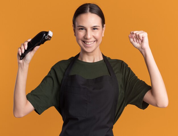 Souriante jeune fille de barbier brune en uniforme garde le poing et tient une tondeuse à cheveux isolée sur un mur orange avec espace de copie