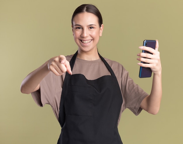 Souriante jeune fille de barbier brune en uniforme pointe vers la caméra et tient le téléphone isolé sur un mur vert olive avec espace de copie