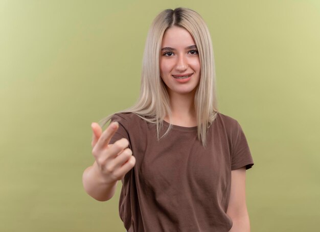 Souriante jeune fille blonde en appareil dentaire faisant venir ici geste sur un espace vert isolé avec copie espace