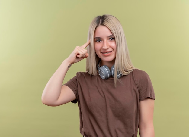 Souriante jeune fille blonde en appareil dentaire portant des écouteurs sur le cou avec le doigt sur le temple sur un espace vert isolé