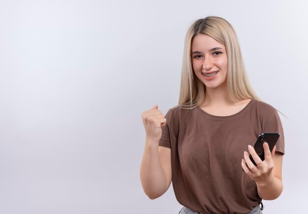 Souriante jeune fille blonde en appareil dentaire tenant un téléphone mobile avec le poing levé sur un espace blanc isolé avec copie