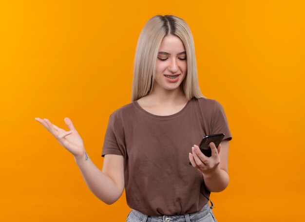 Souriante jeune fille blonde dans un appareil dentaire tenant un téléphone mobile en le regardant montrant une main vide sur un espace orange isolé avec copie espace