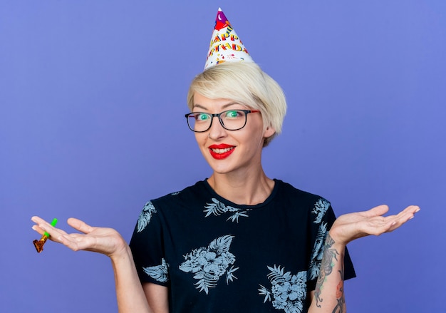 Souriante jeune fille blonde de fête portant des lunettes et une casquette d'anniversaire tenant un ventilateur de fête regardant la caméra montrant les mains vides isolés sur fond violet