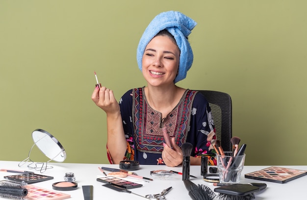 Souriante jeune fille brune aux cheveux enveloppés dans une serviette assise à table avec des outils de maquillage tenant un brillant à lèvres