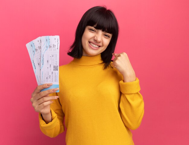 Souriante jeune fille brune caucasienne garde le poing et détient des billets d'avion isolés sur un mur rose avec espace de copie