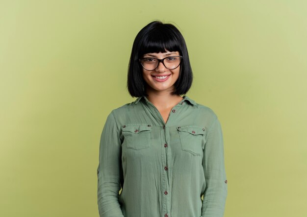 Souriante jeune fille brune caucasienne à lunettes optiques regarde la caméra isolée sur fond vert olive avec espace copie