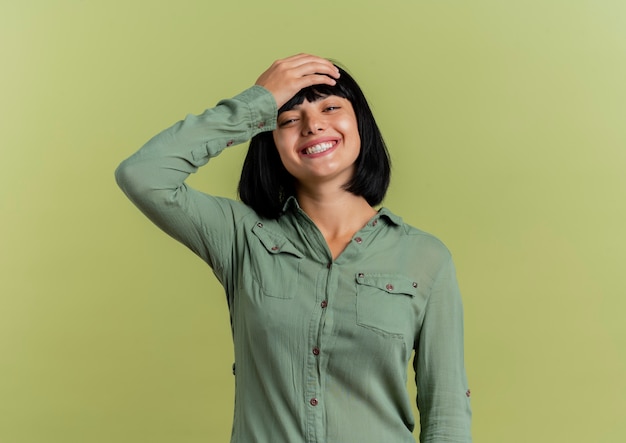 Souriante jeune fille brune caucasienne met la main sur la tête en regardant la caméra isolée sur fond vert olive avec espace copie