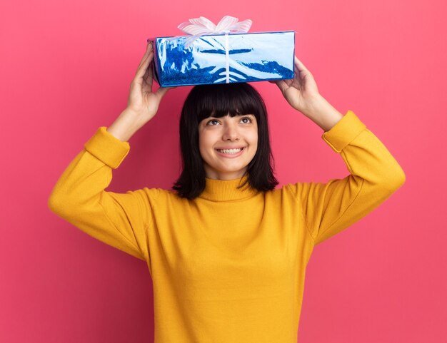 Souriante jeune fille brune caucasienne tient une boîte-cadeau sur la tête rose