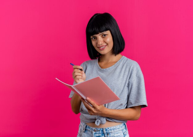 Souriante jeune fille brune caucasienne tient un stylo et un cahier et ressemble