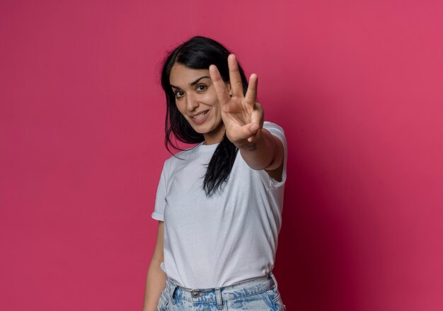 Souriante jeune fille brune caucasienne trois gestes avec la main isolée sur un mur rose