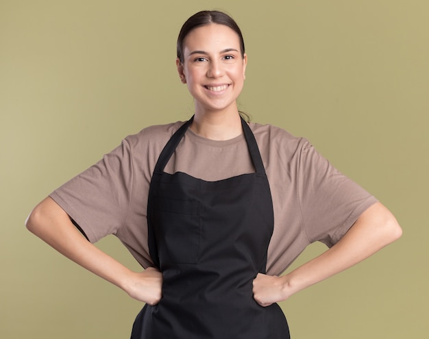 Souriante jeune fille brune de coiffure en uniforme met les poings sur la taille