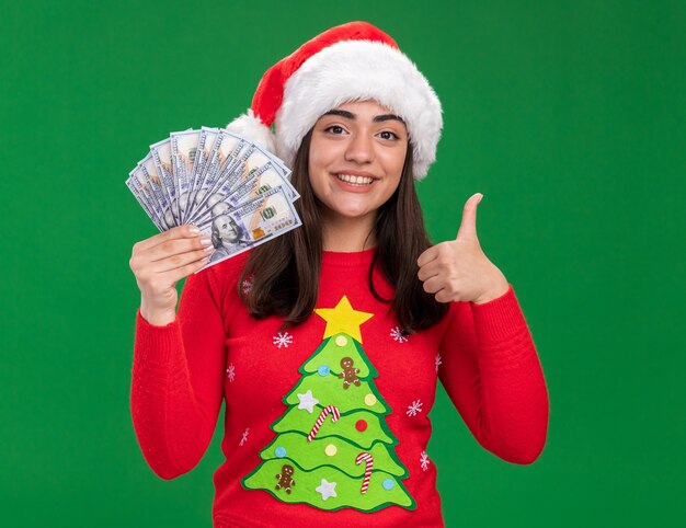 Souriante jeune fille caucasienne avec bonnet de Noel détient de l'argent et les pouces vers le haut isolé sur fond vert avec espace copie