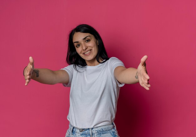 Souriante jeune fille caucasienne brune qui s'étend les mains en faisant semblant de tenir quelque chose d'isolé sur un mur rose