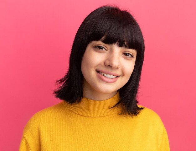 Souriante jeune fille caucasienne brune se tient debout en regardant la caméra isolée sur un mur rose avec espace de copie