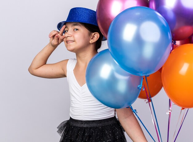 Souriante jeune fille caucasienne avec chapeau de fête bleu tenant des ballons d'hélium et signe de la main de la victoire des gestes isolé sur un mur blanc avec espace copie