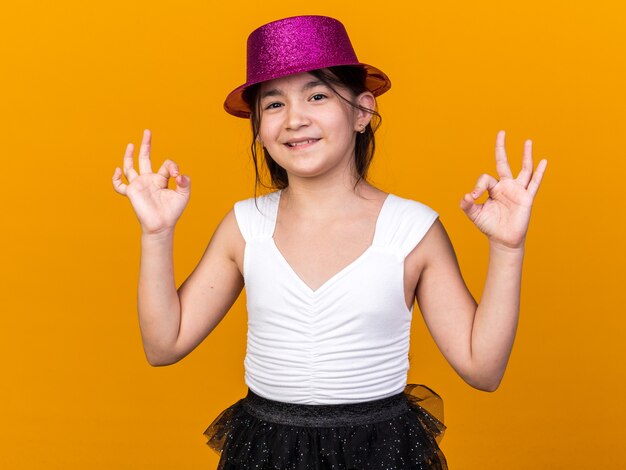 souriante jeune fille caucasienne avec chapeau de fête violet gesticulant signe ok isolé sur mur orange avec espace de copie