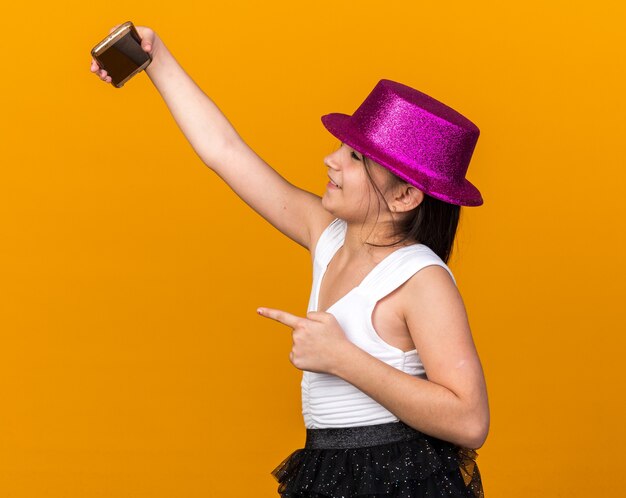 souriante jeune fille caucasienne avec chapeau de fête violet pointant sur le téléphone prenant selfie isolé sur mur orange avec espace de copie