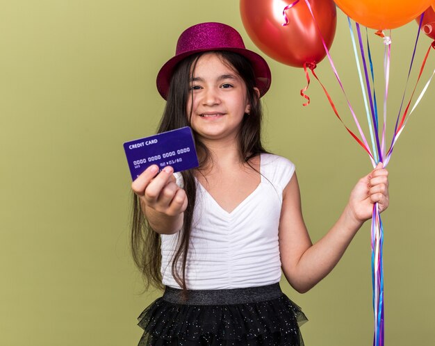 souriante jeune fille caucasienne avec chapeau de fête violet tenant une carte de crédit et des ballons à l'hélium isolés sur un mur vert olive avec espace de copie
