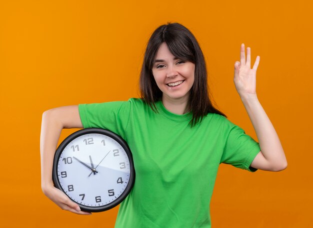Souriante jeune fille caucasienne en chemise verte tient horloge et fait un geste ok sur fond orange isolé