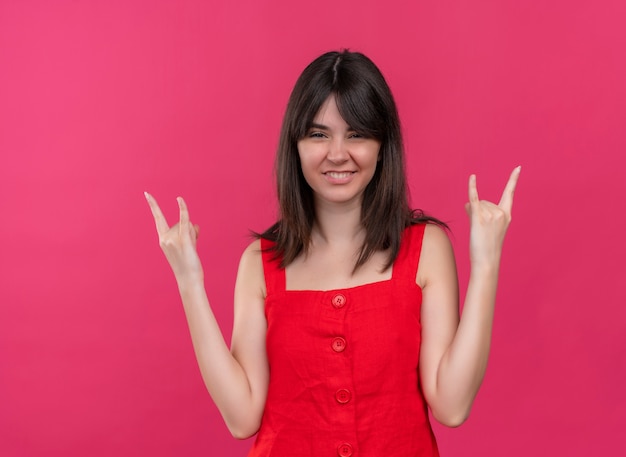 Souriante jeune fille caucasienne fait signe de cornes avec les deux mains sur fond rose isolé