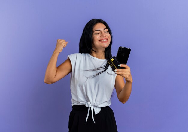 Souriante jeune fille caucasienne garde le poing et détient la carte de crédit du téléphone