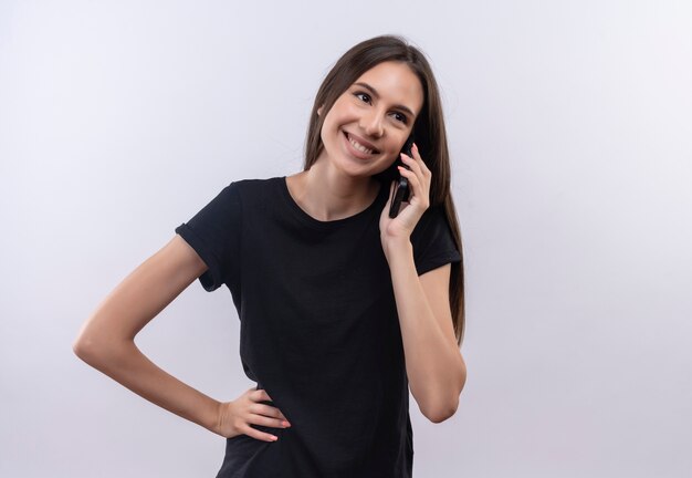 Souriante jeune fille caucasienne portant un t-shirt noir parle au téléphone a mis sa main sur la hanche sur fond blanc isolé