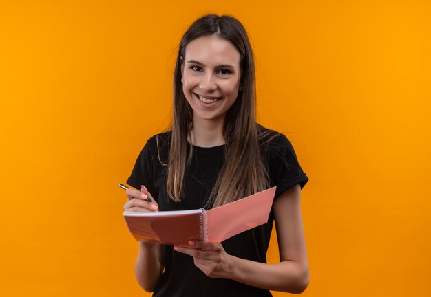 Souriante jeune fille caucasienne portant un t-shirt noir tenant un cahier et un stylo sur fond orange isolé