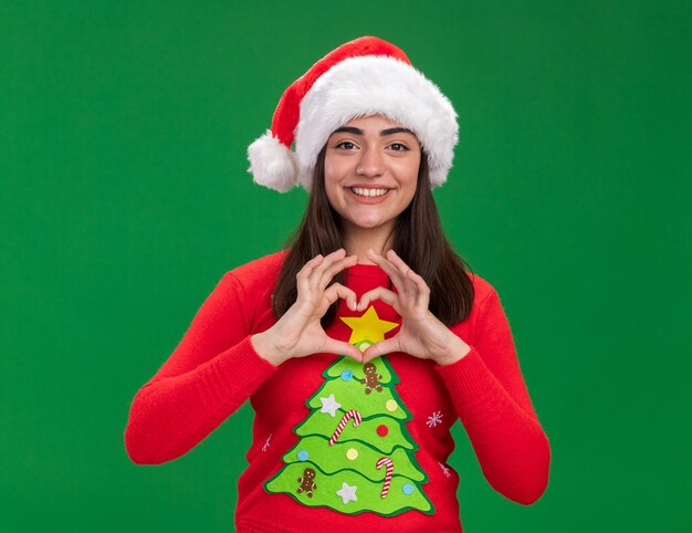 Souriante jeune fille caucasienne avec santa hat gestes signe de coeur isolé sur fond vert avec espace copie