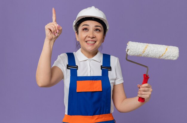 Souriante jeune fille de constructeur asiatique avec un casque de sécurité blanc tenant un rouleau à peinture et pointant vers le haut