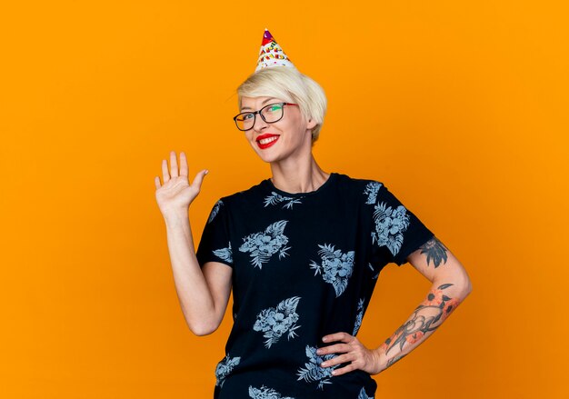 Souriante jeune fille de fête blonde portant des lunettes et une casquette d'anniversaire regardant la caméra en gardant la main sur la taille faisant salut caméra geste isolé sur fond orange avec espace copie