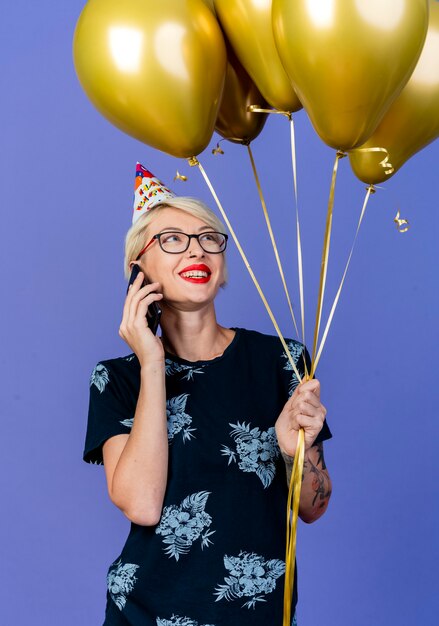 Souriante jeune fille de fête blonde portant des lunettes et une casquette d'anniversaire tenant et regardant des ballons parler au téléphone isolé sur fond violet