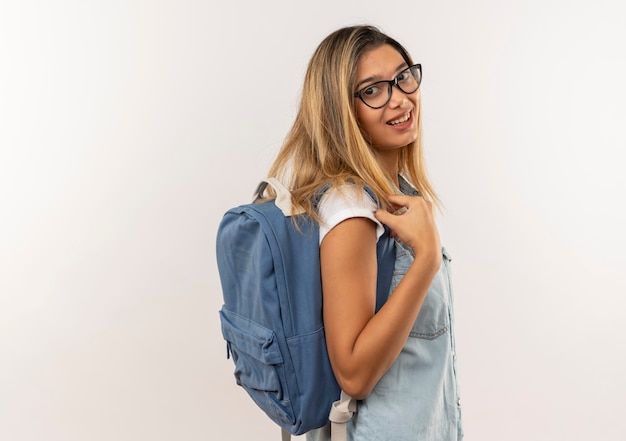 Souriante jeune fille jolie étudiante portant des lunettes et sac à dos debout en vue de profil à l'avant isolé sur un mur blanc