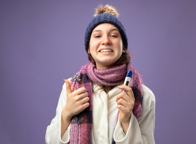 Souriante jeune fille malade portant une robe blanche et un chapeau d'hiver avec écharpe tenant un thermomètre montrant le pouce vers le haut isolé sur violet