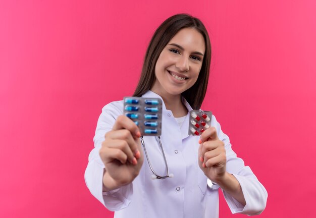 Souriante jeune fille médecin portant une robe médicale stéthoscope tenant des pilules sur fond rose isolé