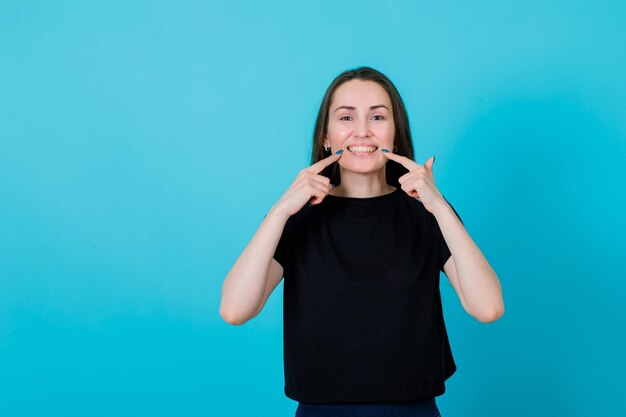 Souriante jeune fille montre son sourire avec les index sur fond bleu