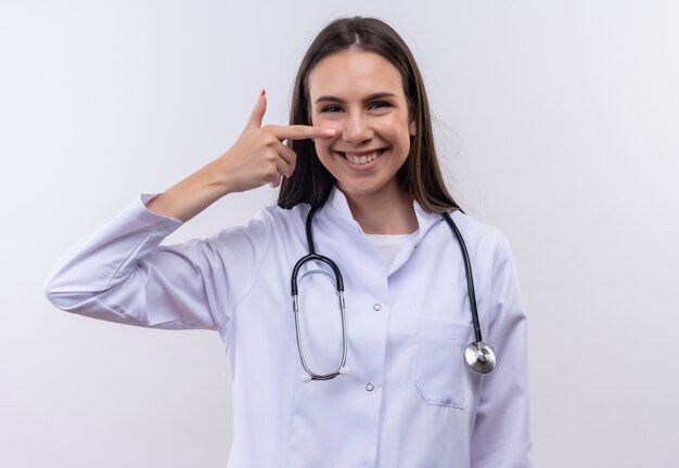 Souriante jeune fille portant une robe médicale stéthoscope pointe du doigt sur le nez sur fond blanc isolé