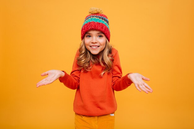 Souriante jeune fille en pull et chapeau hausse les épaules et regardant la caméra sur orange